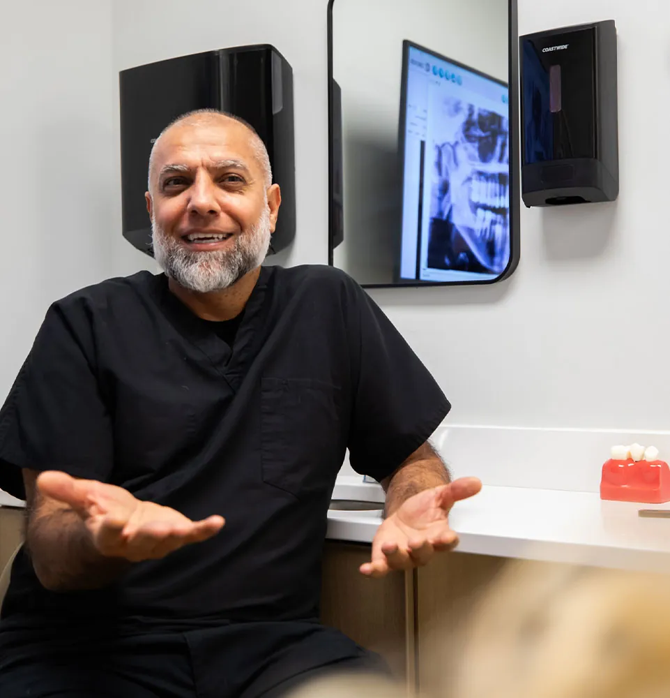Dentist in black scrubs engaging with a patient, discussing dental care in a modern clinic setting, with dental x-rays displayed on a screen and dental tools visible.